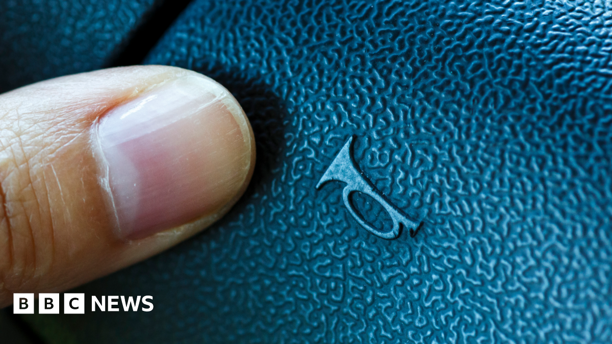 A close-up of someone's thumb on the dashboard button used to sound the horn. The black plastic button, which has a faux-leather pattern, has the symbol of a trumpet-like horn on it.
