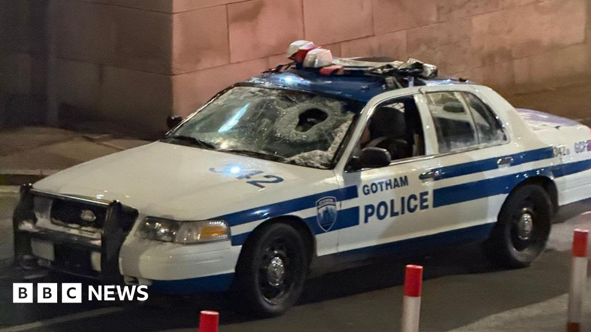 A blue and white liveried police car emblazoned with the sign of Gotham Police. the front windscreen is shattered with a hole in the glass.