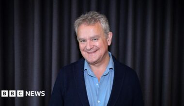 The image shows a man smiling at the camera; he is wearing a light blue buttoned top and a navy blue blazer.