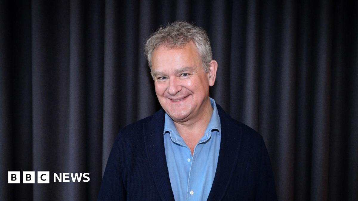 The image shows a man smiling at the camera; he is wearing a light blue buttoned top and a navy blue blazer.