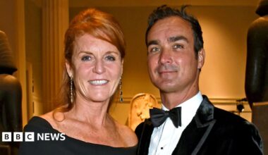 Sarah Ferguson in black dress with her hair up stands next to Manuel Fernandez who wears a black velvet jacket, white shirt and bow tie. They are both smiling at the camera.