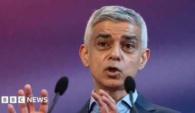 Sadiq Khan is pictured from the shoulders up giving a speech. Two microphones are visible in the foreground and he stands in front of a blurred purple background.