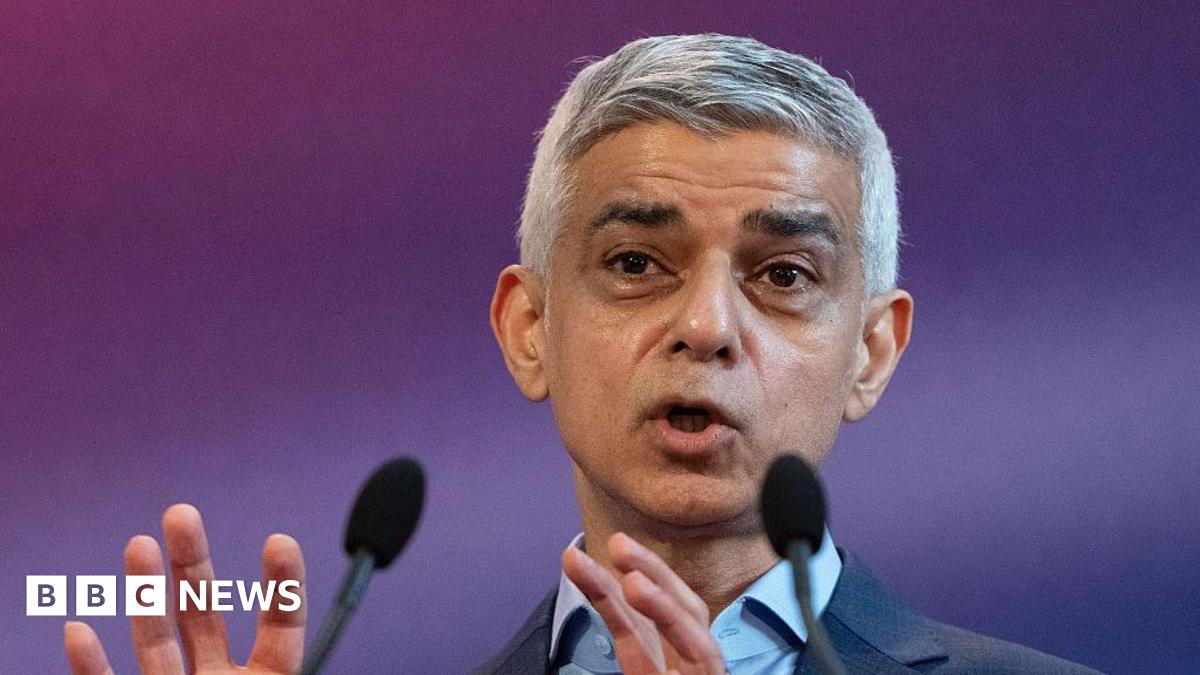 Sadiq Khan is pictured from the shoulders up giving a speech. Two microphones are visible in the foreground and he stands in front of a blurred purple background.