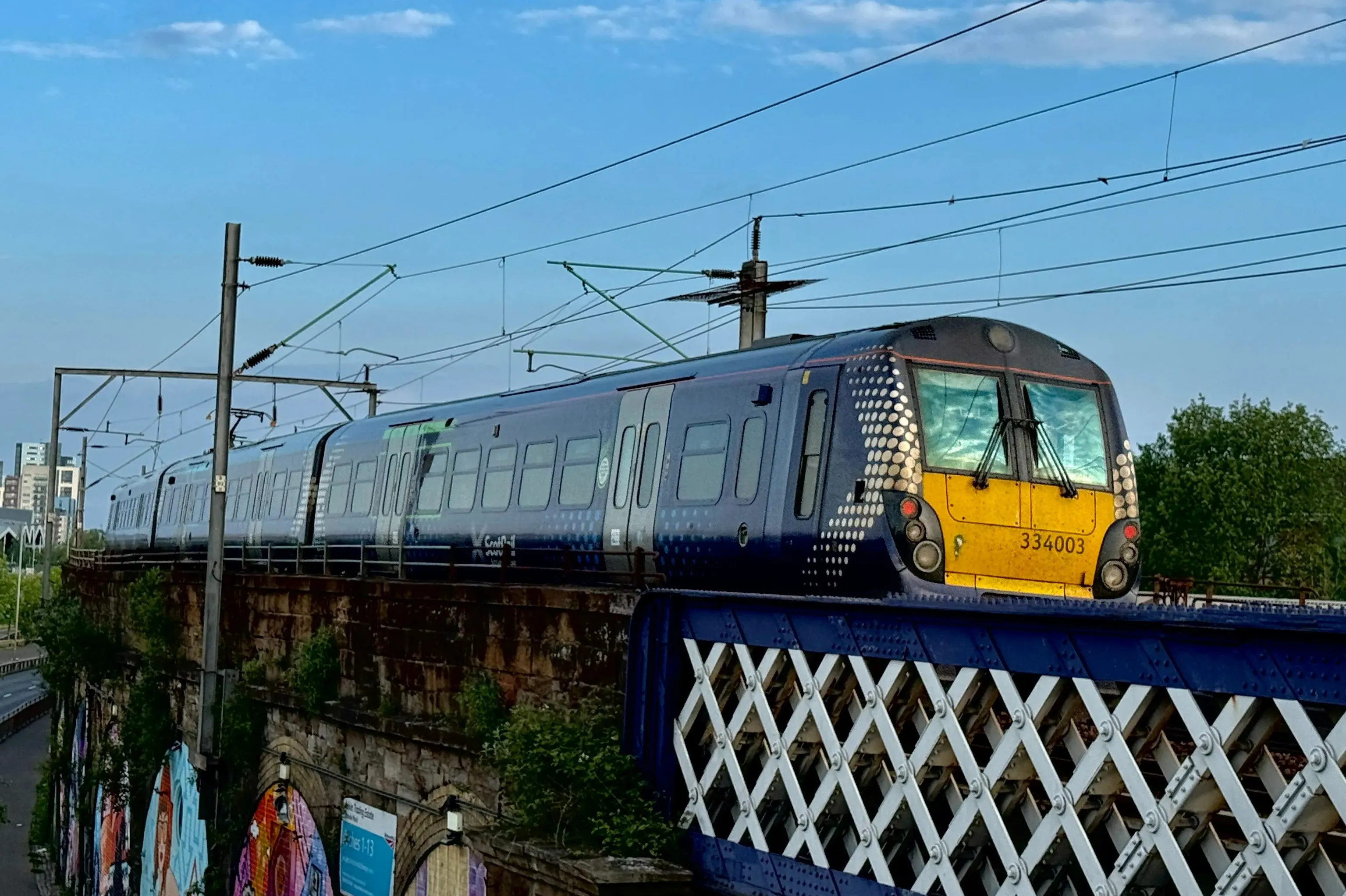 A blue and yellow train crosses a metal bridge with a crisscross pattern, carrying overhead power lines, under a blue sky with scattered clouds.