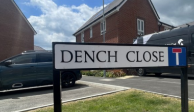 The Dench Close street sign fixed into the grass kerb side as you enter Dench Close with a van, a car and red-brick houses in the background.