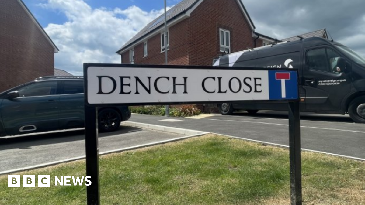 The Dench Close street sign fixed into the grass kerb side as you enter Dench Close with a van, a car and red-brick houses in the background.