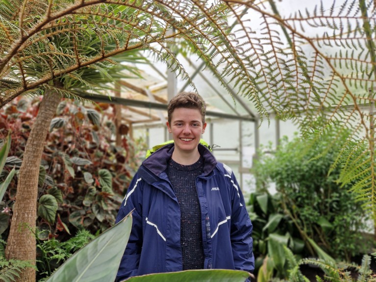 Daniella Crowley smiling in a greenhouse at Treborth surrounded by treeferns and other plants.