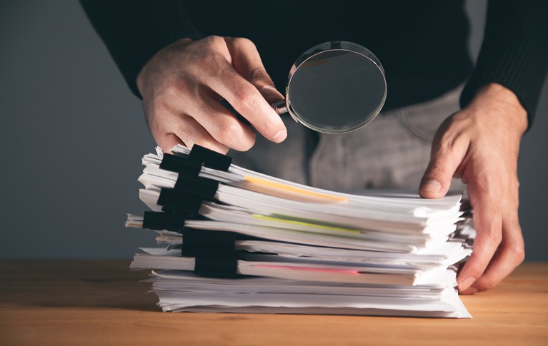 A close-up view, showing hands of a person holding a magnifying glass over a pile of documents on a tabletop.