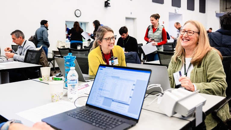 Rosemarie Wilton at a hackathon event, seated with a laptop and laughing with a female colleague.