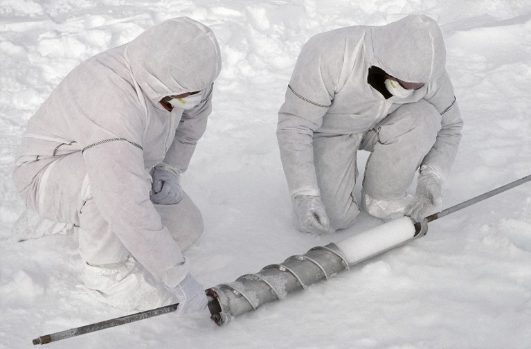Two researchers wearing white boiler suits extract an ice core from the drill bit, Antarctica.
