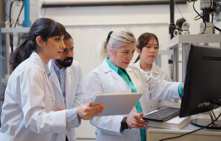 Four scientists in white lab coats collaborating at a computer in a laboratory.