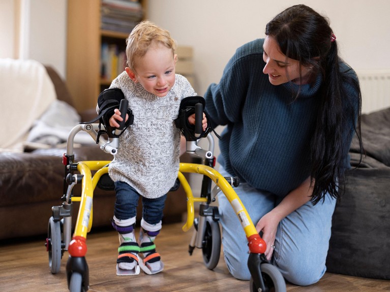 Mother helping her young child with a neuromuscular condition to walk using a four wheeled walker.