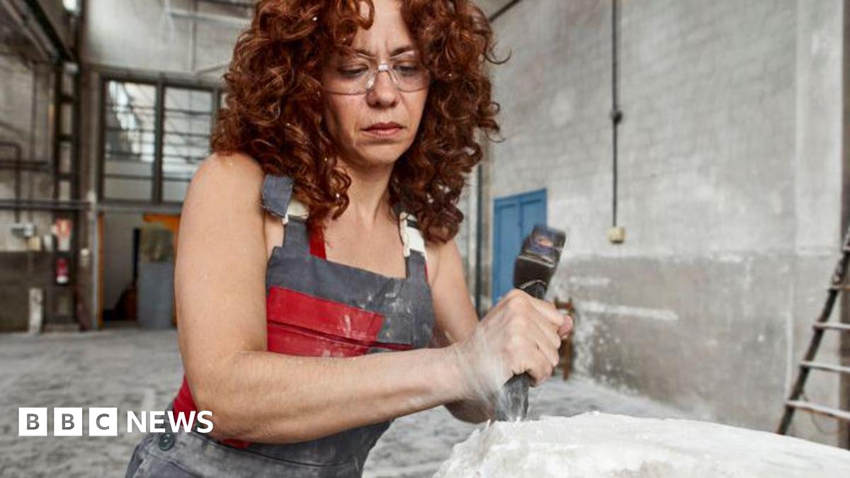 A person wearing glasses and a protective apron stands in a large indoor workshop, using a hammer and chisel to shape a block of pale stone. Fine dust is visible and the surface of the stone is rough and partially formed. The background shows a high‑ceilinged industrial space with white walls, exposed fixtures, and a ladder leaning against the wall.