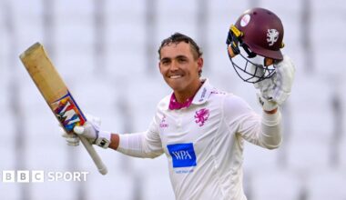 Josh Thomas holds his bat in the air in his right hand and his helmet in his left as he smiles after scoring a century against Yorkshire