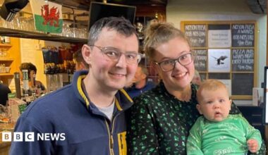 On the right is a man with dark hair, dark eyes and glasses. He's smiling at the camera. He's wearing a navy and yellow jumper. Next to him is a woman with light hair and eyes. She's wearing glasses and is smiling at the camera. She has a green and black top on. She's holding a baby which has blonde hair and blue eyes. He's wearing a green t-shirt and blue trousers.