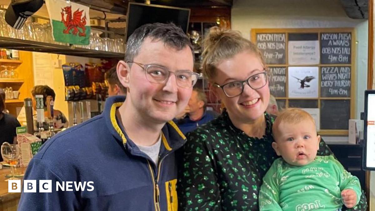 On the right is a man with dark hair, dark eyes and glasses. He's smiling at the camera. He's wearing a navy and yellow jumper. Next to him is a woman with light hair and eyes. She's wearing glasses and is smiling at the camera. She has a green and black top on. She's holding a baby which has blonde hair and blue eyes. He's wearing a green t-shirt and blue trousers.