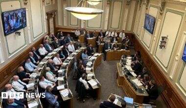 Looking down into a council chambers, circular rows of desks with people sat in smart clothes. In front of them a smaller desk with five people sat facing them. Screens are on the walls of the room and there are overhanging lights.