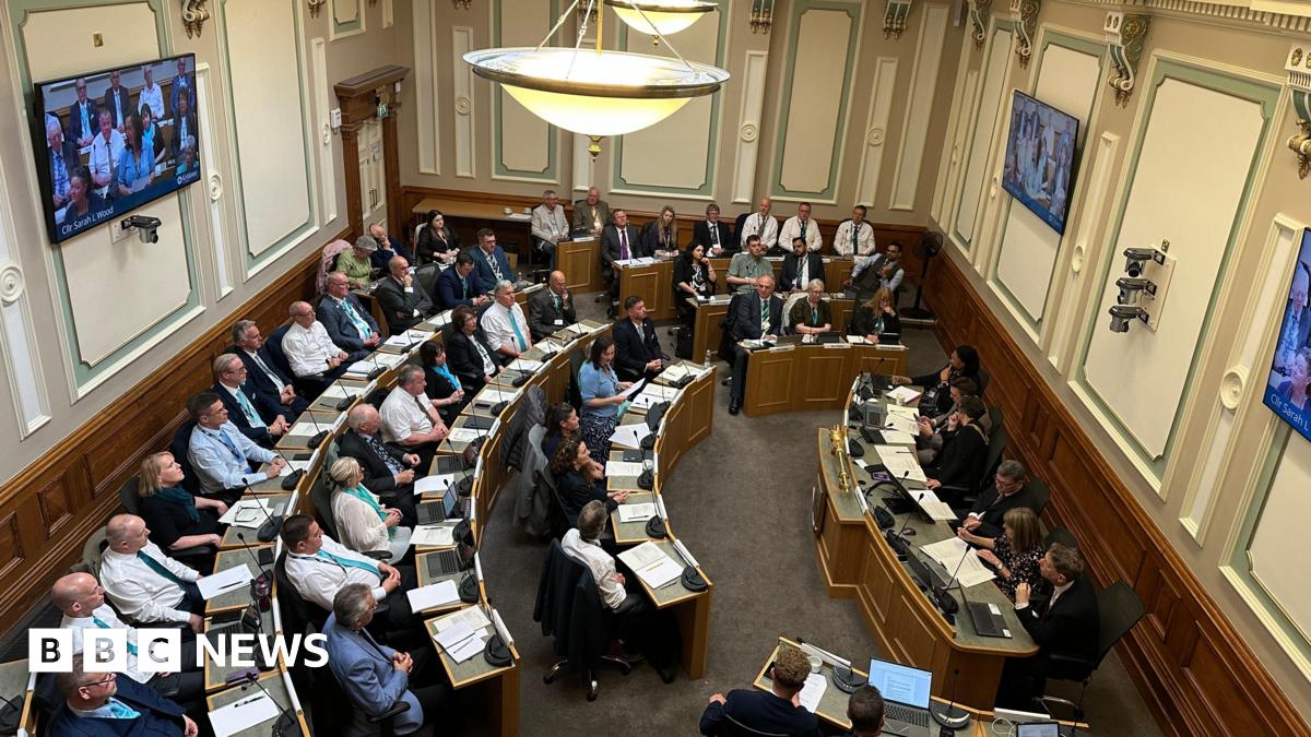 Looking down into a council chambers, circular rows of desks with people sat in smart clothes. In front of them a smaller desk with five people sat facing them. Screens are on the walls of the room and there are overhanging lights.