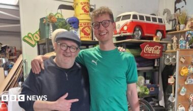 Two men in glasses, each with an arm around the shoulder of the other. They are both smiling, the older man is wearing a flat cap
