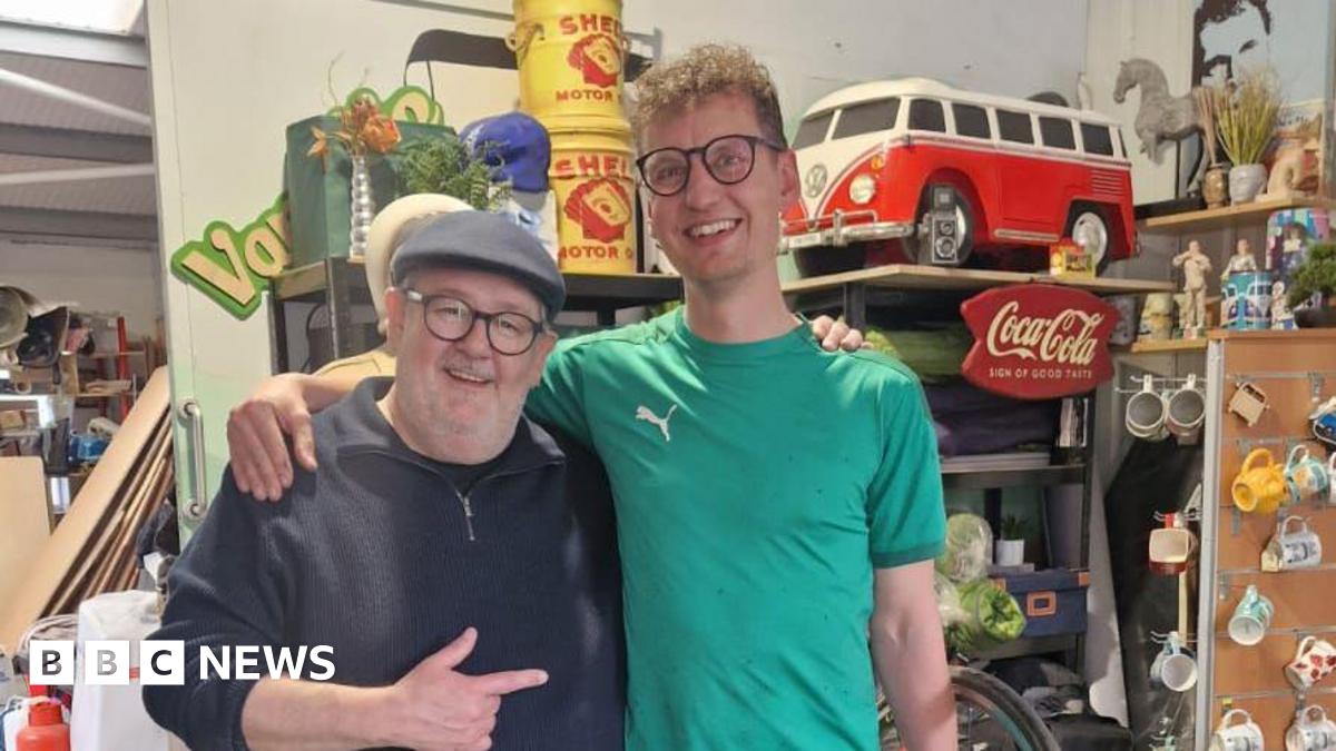 Two men in glasses, each with an arm around the shoulder of the other. They are both smiling, the older man is wearing a flat cap