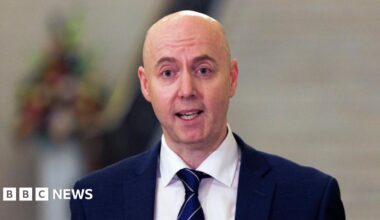 Jon Burrows is wearing a white shirt with navy blazer and tie. The background is blurry.