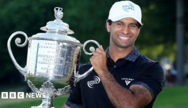 Rai holds a massive trophy and is smiling at the camera. He is wearing a white cap and navy top.