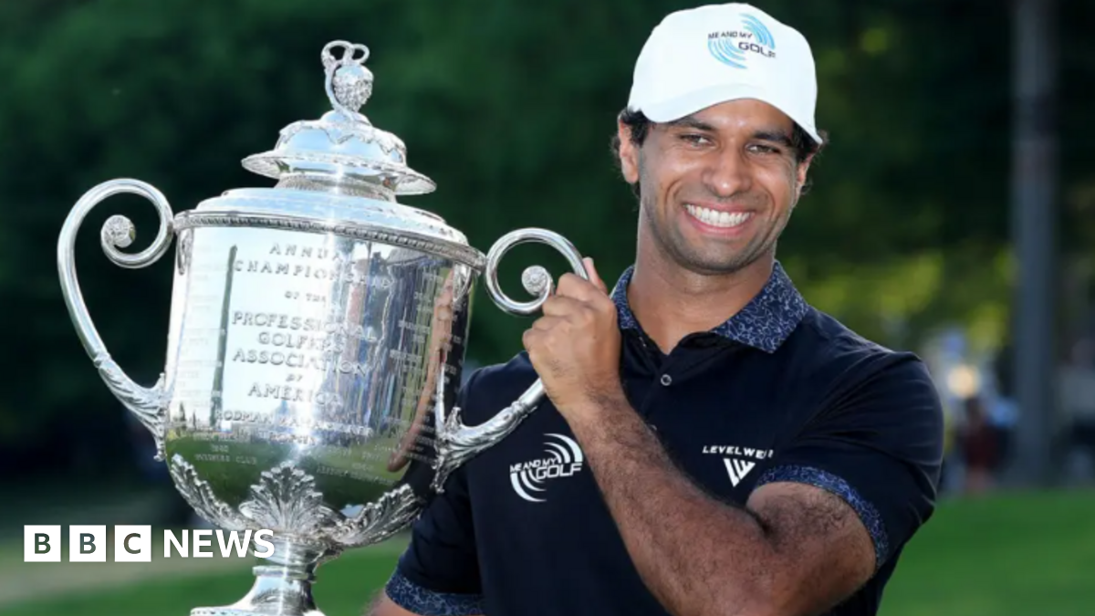 Rai holds a massive trophy and is smiling at the camera. He is wearing a white cap and navy top.