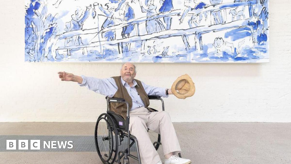 Quentin Blake, sat in a wheelchair, wearing a light blue shirt and brown waist jacket and holding a tan hat with his arms outstretched under a large blue and white mural.