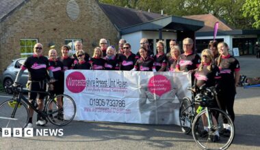 About 20 cyclists all wearing black cycling outfits stand with their bikes and hold up a large white banner for the Worcestershire Breast Unit Haven. The tag line "everybody knows somebody" is also printed on the banner with a phone number and website for the service.