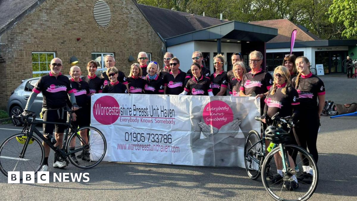 About 20 cyclists all wearing black cycling outfits stand with their bikes and hold up a large white banner for the Worcestershire Breast Unit Haven. The tag line "everybody knows somebody" is also printed on the banner with a phone number and website for the service.