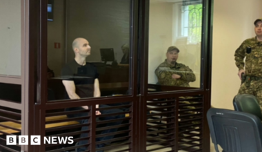 A man in a black t-shirt sits in a witness box guarded by soldiers