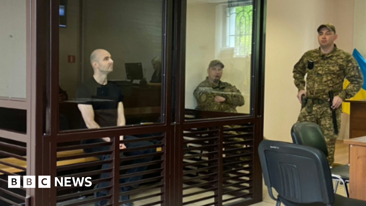 A man in a black t-shirt sits in a witness box guarded by soldiers