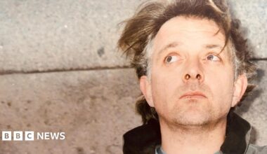 Comedian and actor Rik Mayall, standing with his back against a concrete wall. His hair, mid-brown with grey patches on the sides, is unkempt and he has a five o'clock shadow. He is looking up and behind the camera.