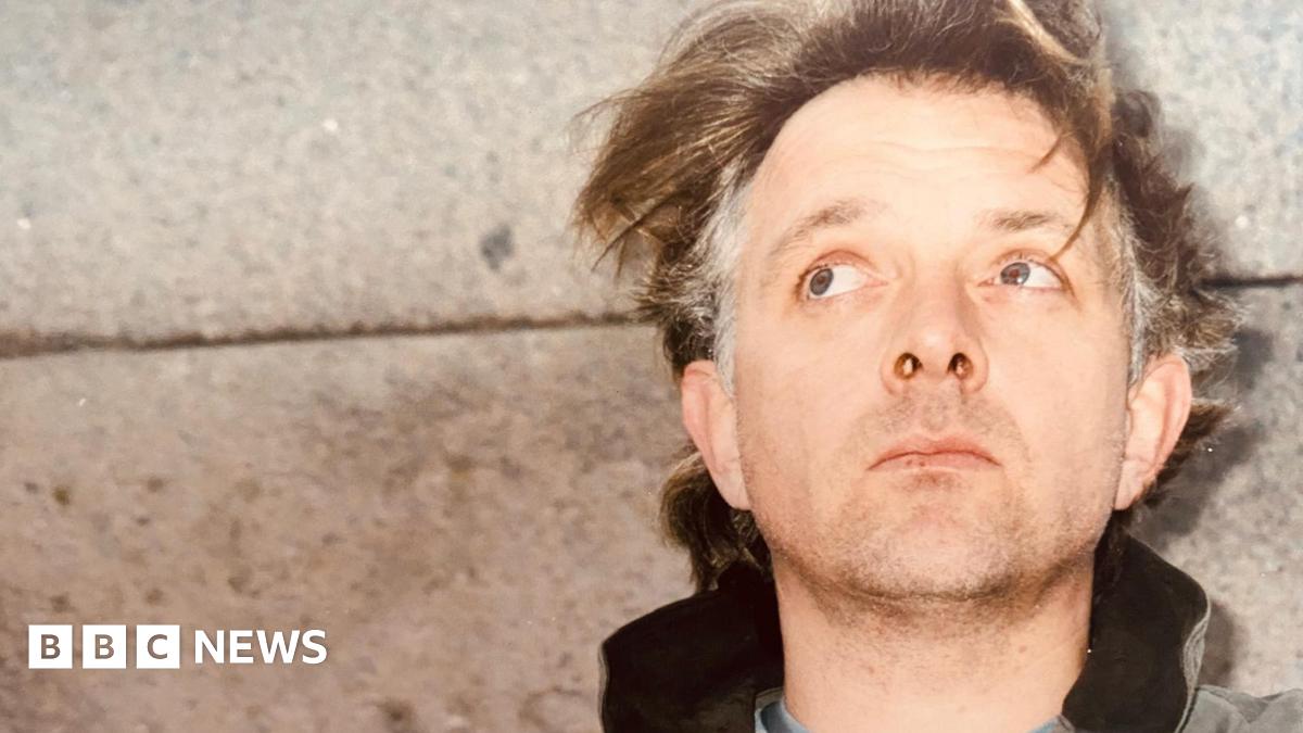 Comedian and actor Rik Mayall, standing with his back against a concrete wall. His hair, mid-brown with grey patches on the sides, is unkempt and he has a five o'clock shadow. He is looking up and behind the camera.