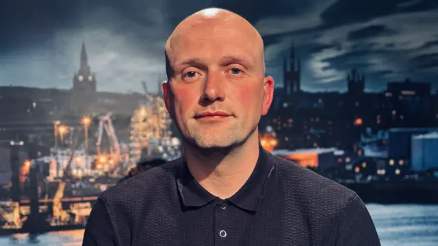 Stephen Flynn, a bald man wearing a black polo shirt.