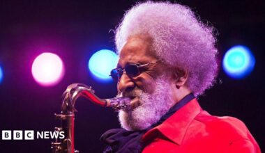 Sonny Rollins plays saxophone while wearing a red jacket on stage under blue and pink lights.