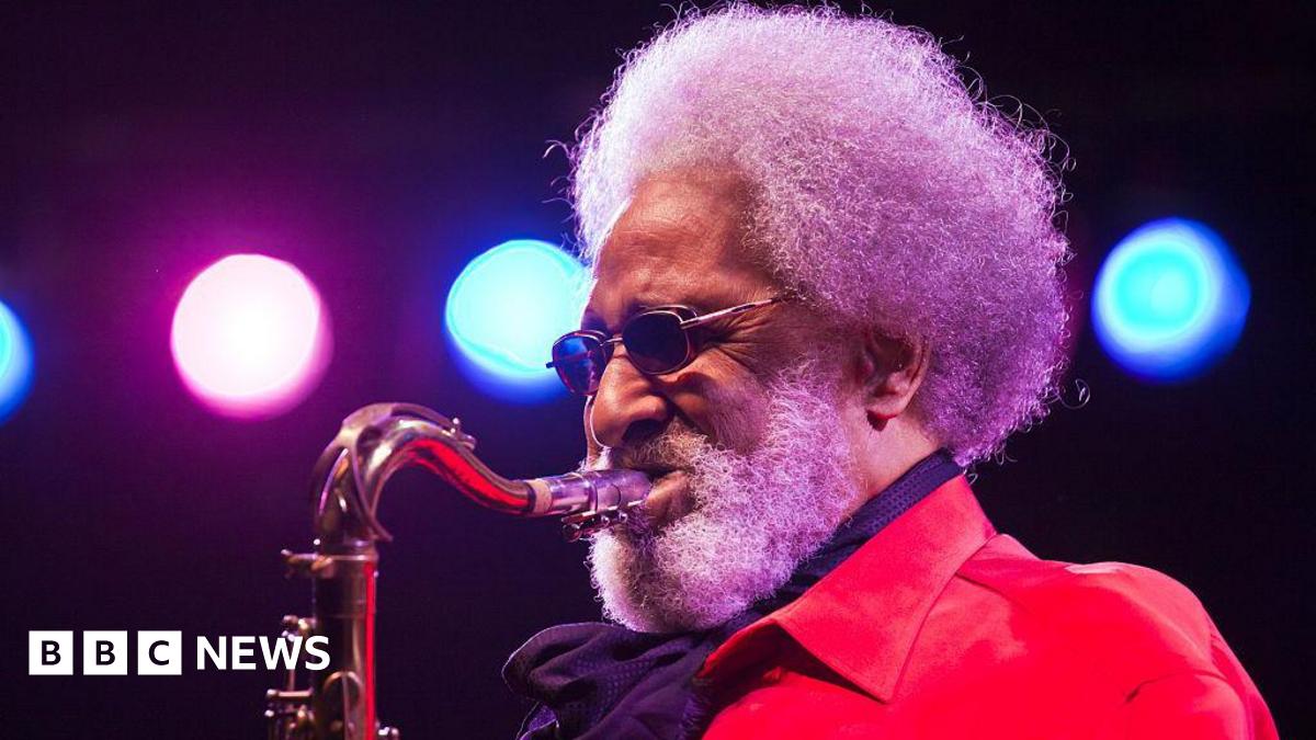 Sonny Rollins plays saxophone while wearing a red jacket on stage under blue and pink lights.