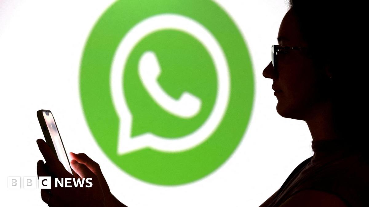A teenager poses for a photo while holding a smartphone in front of a Whatsapp logo