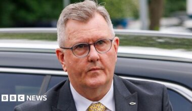 A man who has just come out of a car with a neutral expression on his face. He is wearing glasses and has short cropped, slightly thinning greyish hair. He is wearing a dark blue suit jacket, white shirt and yellow patterned tie.