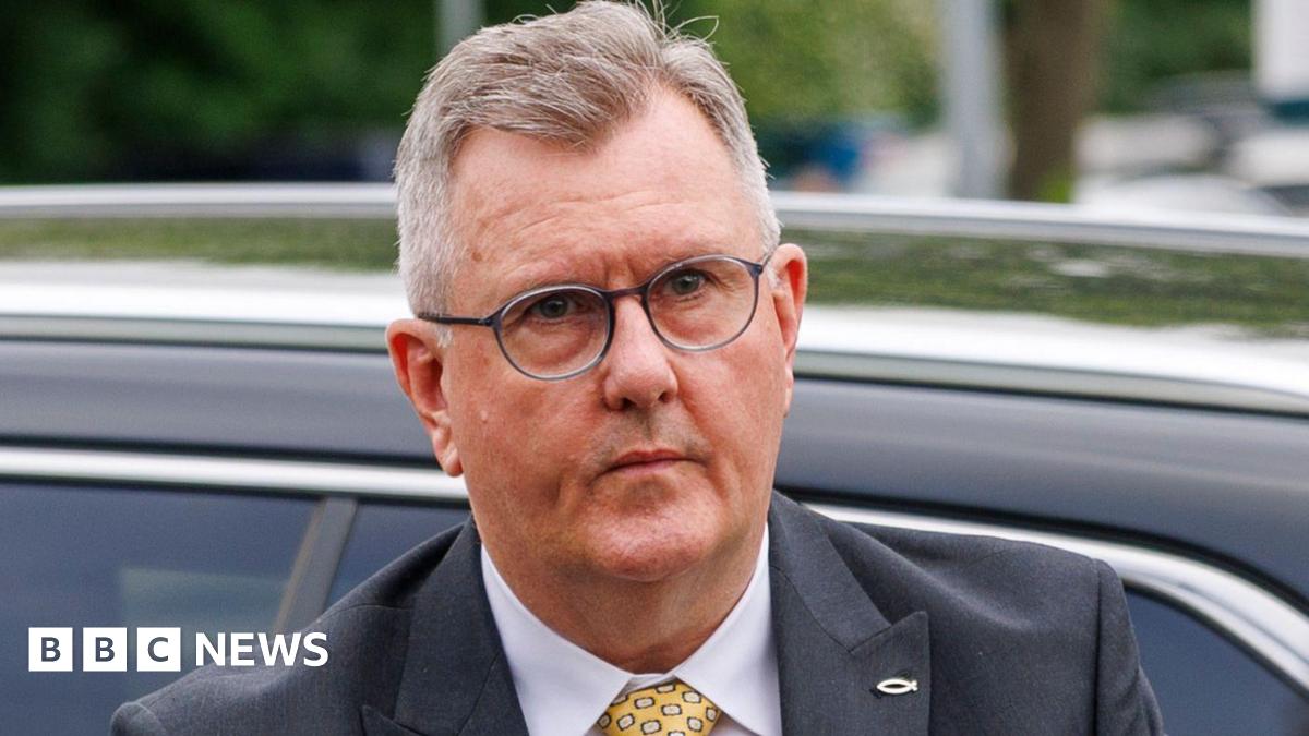 A man who has just come out of a car with a neutral expression on his face. He is wearing glasses and has short cropped, slightly thinning greyish hair. He is wearing a dark blue suit jacket, white shirt and yellow patterned tie.