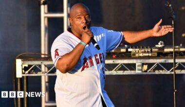 Young MC performs onstage. He is wearing a white Los Angeles jersey with red writing and is singing into a hand-held microphone