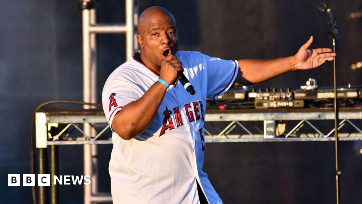 Young MC performs onstage. He is wearing a white Los Angeles jersey with red writing and is singing into a hand-held microphone