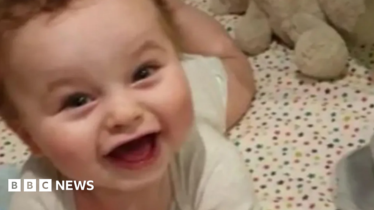 Baby Preston Davey smiles at the camera dressed in a white vest and appears to be lying in a cot with star bedsheet cover