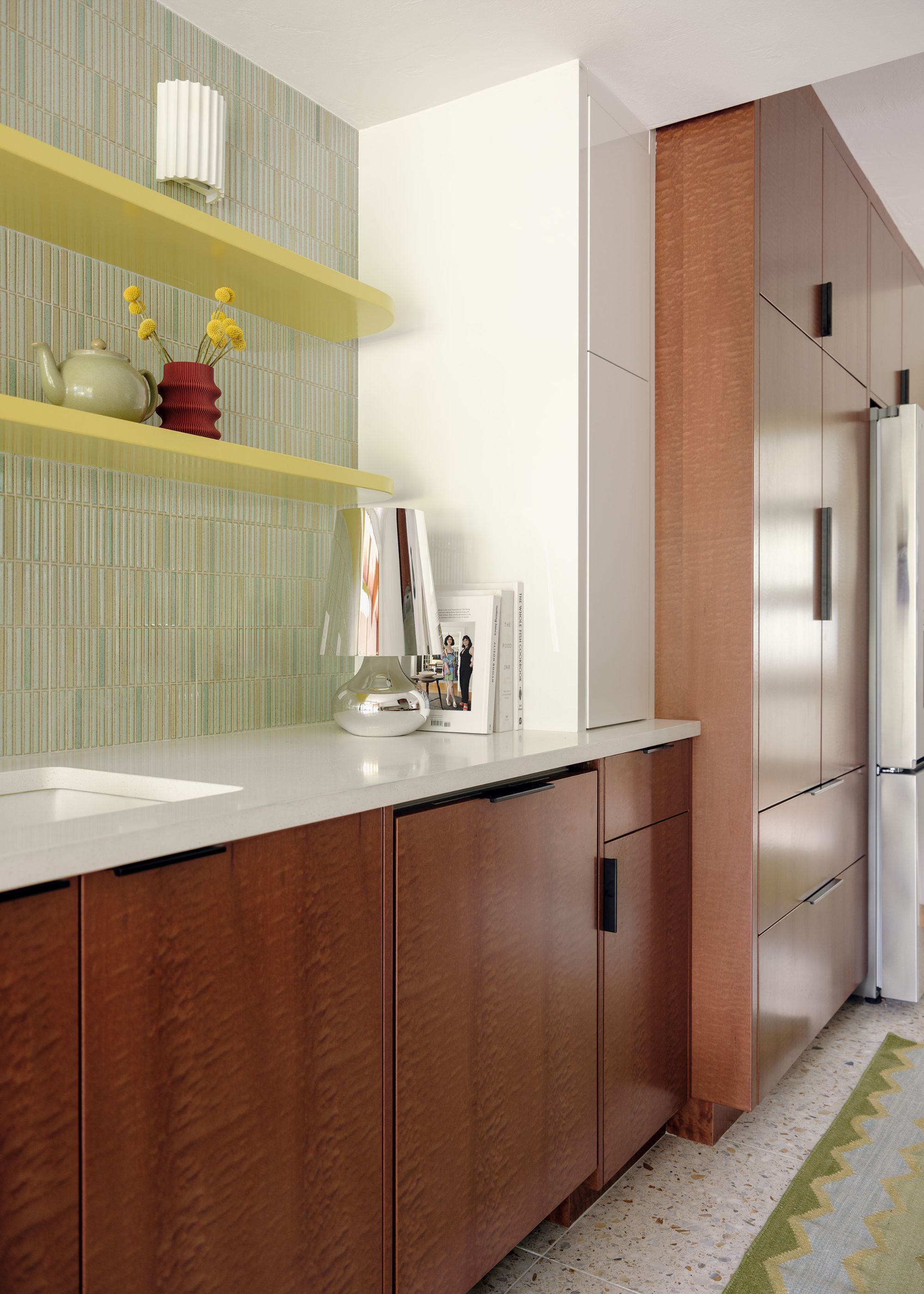 A modern, sleek kitchen with warm wood cabinets, white walls, a mint green tiled accent wall, and lemon yellow shelves.