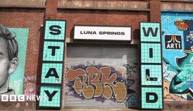 A grey metal shutter with orange graffiti is pulled over the entrance to Luna Springs. Murals are painted on the red brick wall around its entrance. Signs either side of the entrance say 'Stay Wild' in blue, with a white 'Luna Springs' sign above.
