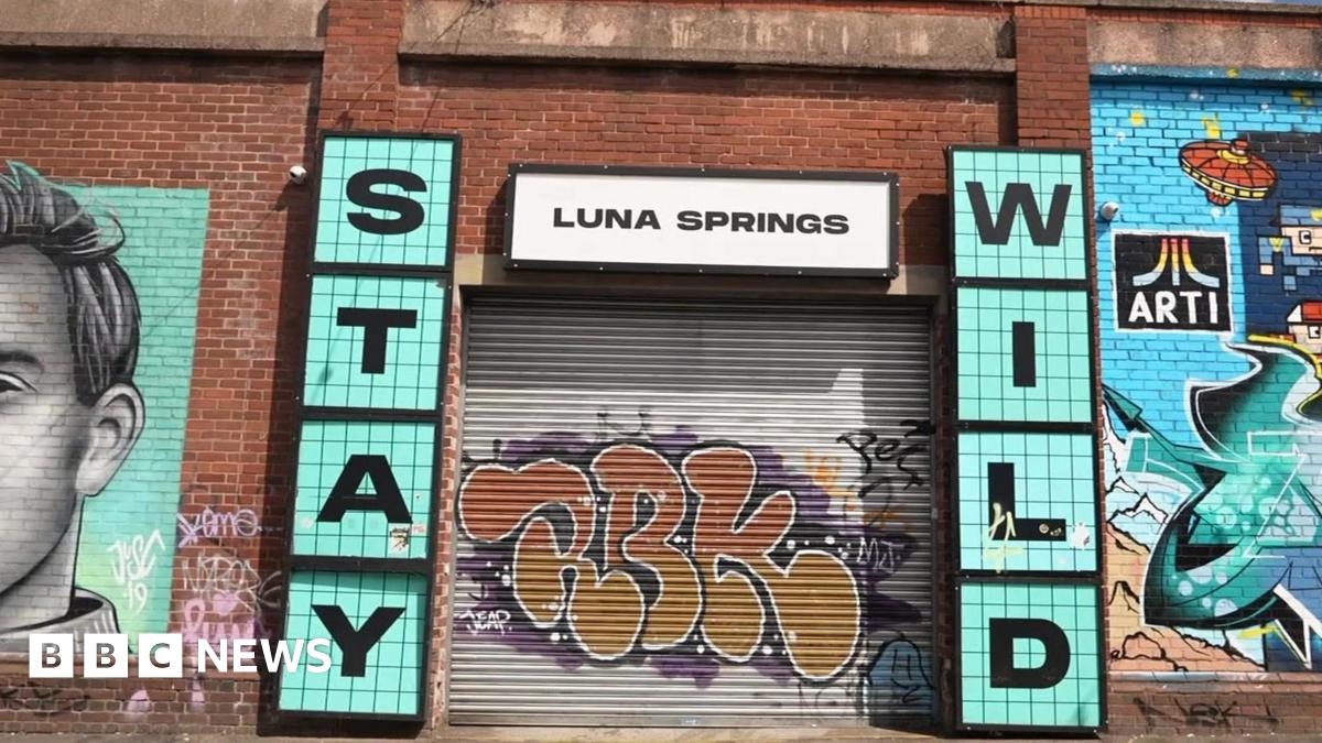 A grey metal shutter with orange graffiti is pulled over the entrance to Luna Springs. Murals are painted on the red brick wall around its entrance. Signs either side of the entrance say 'Stay Wild' in blue, with a white 'Luna Springs' sign above.