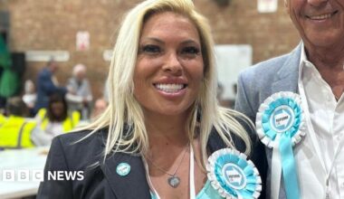 Emma Beck, 47, who has blonde hair, is wearing a turquoise top under a grey blazer, smiles at the camera in a gymnasium. She has a turquoise and white Reform UK party rosette pinned to her lapel.