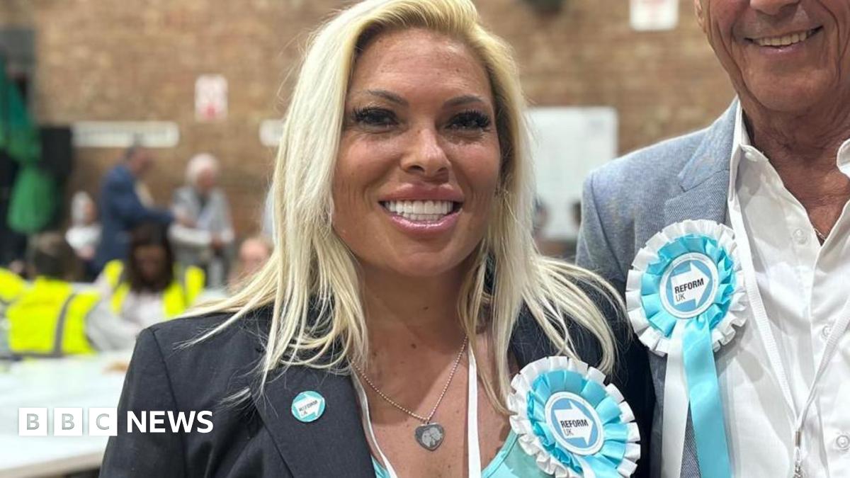 Emma Beck, 47, who has blonde hair, is wearing a turquoise top under a grey blazer, smiles at the camera in a gymnasium. She has a turquoise and white Reform UK party rosette pinned to her lapel.