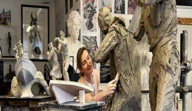 Amy Goodman is pictured in her workshop, using a tool on a statue in progress. Near her are figurines of two men on the prow of a boat, showing how part of the Coastal Forces memorial will look. Photographs of Royal Navy Wrens are positioned in front of her.