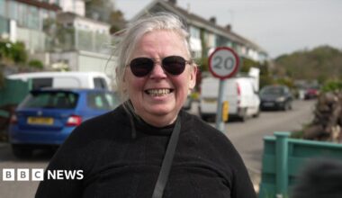 A woman standing on a street. She is wearing sunglasses and is smiling into the camera. Parked cars and houses are behind her.